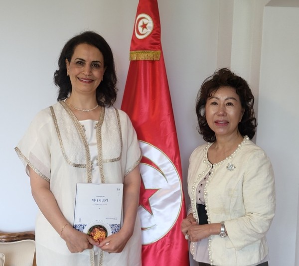 Madam Malika Khadhri El Abed (left), spouse of the Ambassador of Tunisia, and Vice Chairperson Joy Cho of The Korea Post take a picture with the national flag of Tunisia between them.