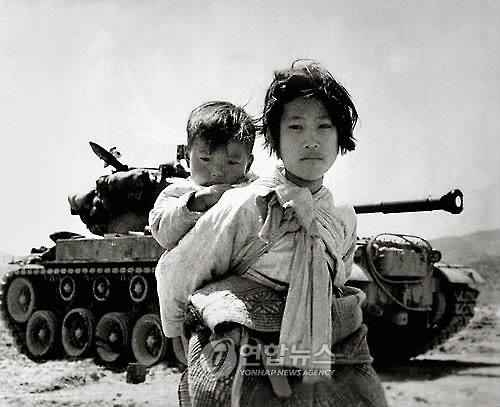 A little Korean girl with her brother on her back leaving her home with refugees to avoid North Korean Invasion Forces during the Korean War.